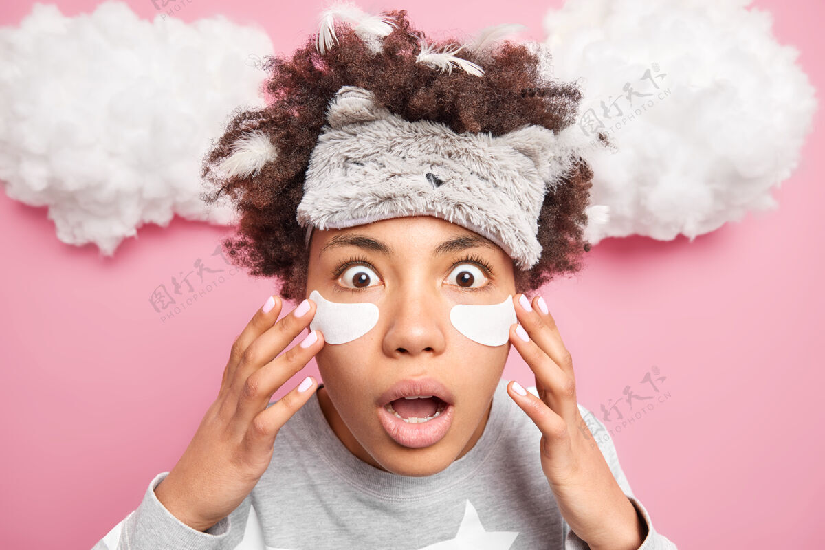 人类特写镜头中 一位表情激动的非裔美国妇女在眼睛下面贴上眼罩 盯着镜头 戴着柔软的眼罩 在粉色的云层上做着护肤动作醒来成人睡衣