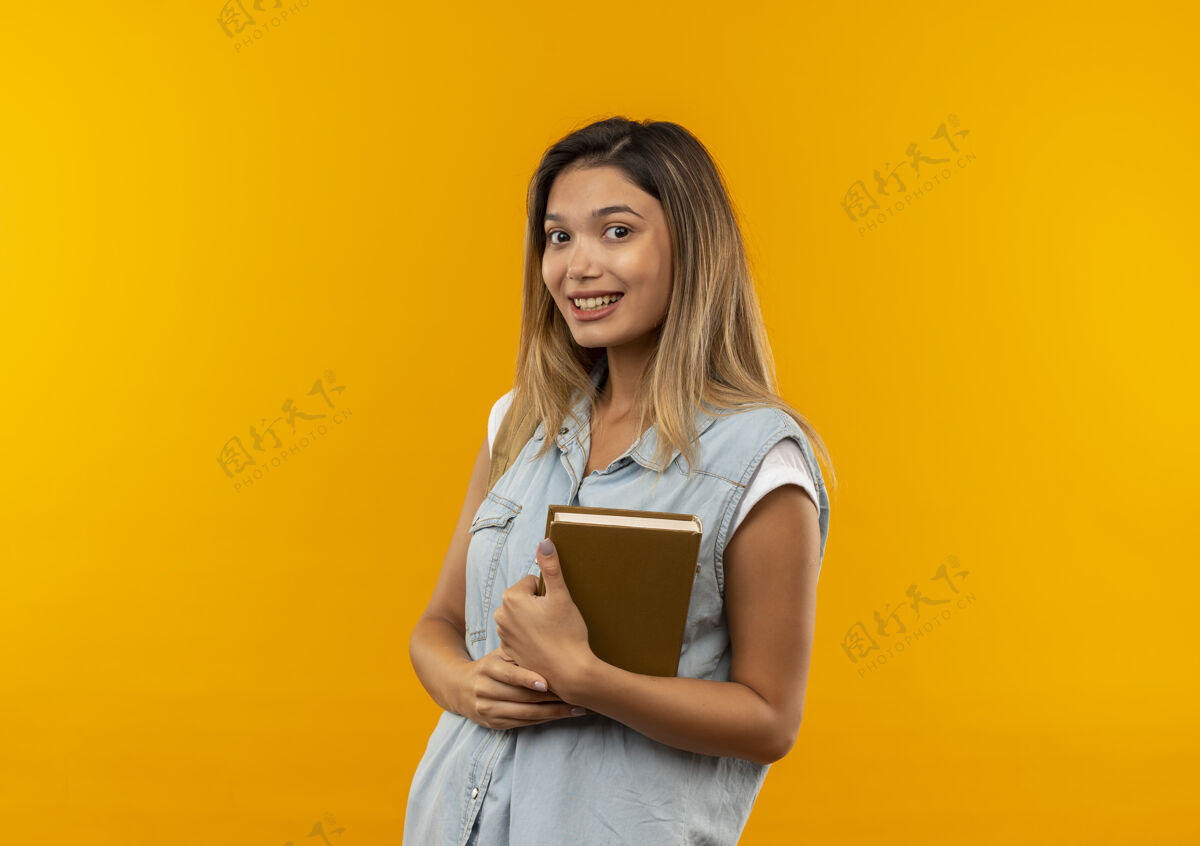 人微笑着的年轻漂亮的学生女孩 背着书包 把书孤立在橙色的墙上公民包橙色