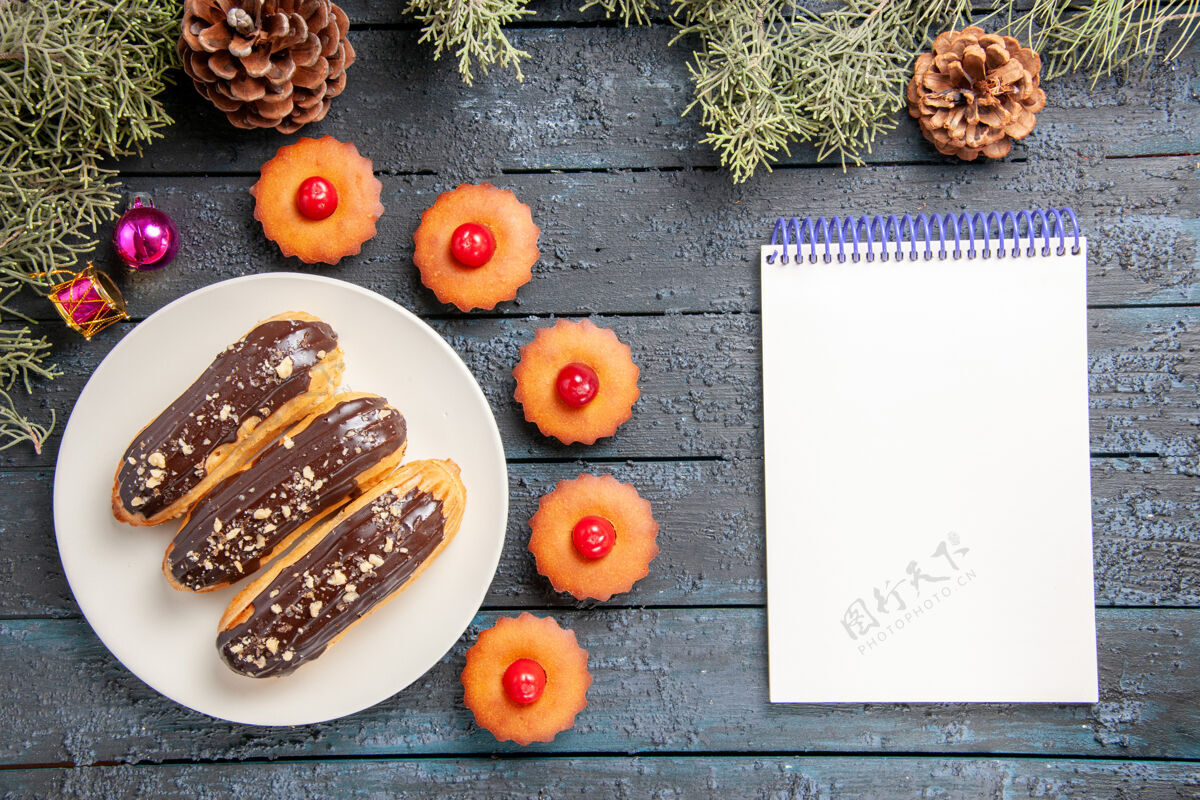 桌子俯视图白色椭圆形平板冷杉树枝上的巧克力蛋糕圣诞玩具樱桃纸杯蛋糕和黑木桌上的笔记本甜点冷杉樱桃