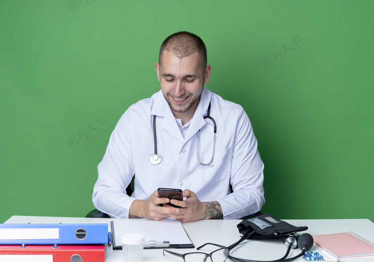 表情面带微笑的年轻男医生穿着医用长袍和听诊器坐在办公桌旁 手里拿着工作工具 看着隔离在绿墙上的手机绿色电话人