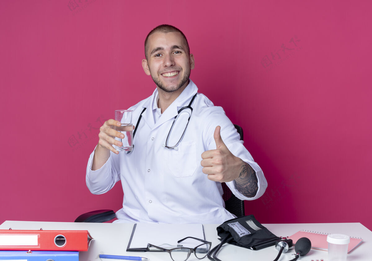 表演面带微笑的年轻男医生穿着医用长袍 手持听诊器坐在办公桌旁 手里拿着一杯水 在粉红色的墙上孤立地竖起大拇指抱着坐着医疗