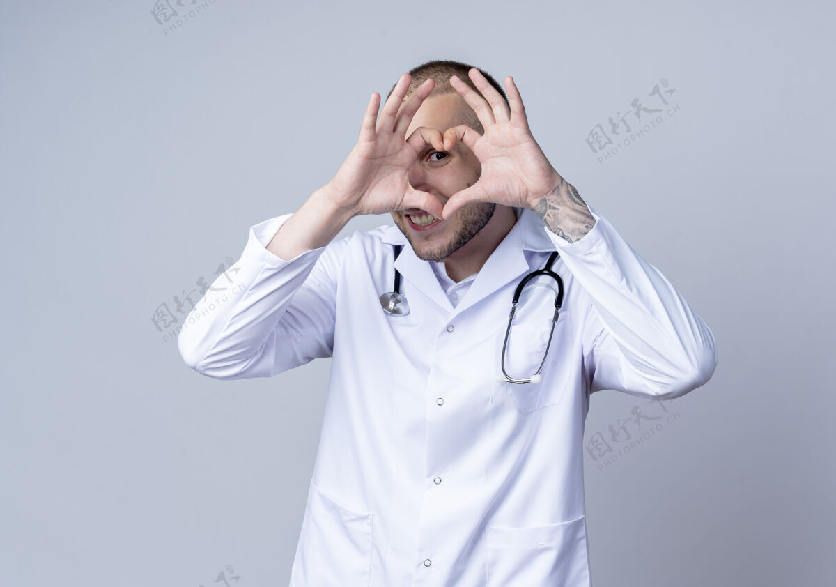 脸面带微笑的年轻男医生 身穿医用长袍 脖子上戴着听诊器 做着心脏信号 隔着隔离在白墙上的门看着前方衣服人听诊器