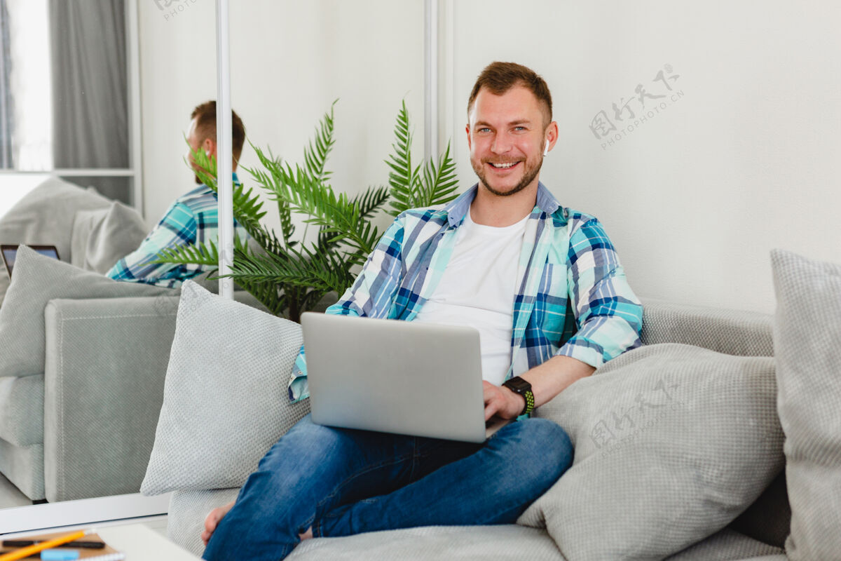 在家工作穿着衬衫的微笑男人在家里的沙发上放松地坐在桌子旁 在家里用笔记本电脑上网成人沙发使用