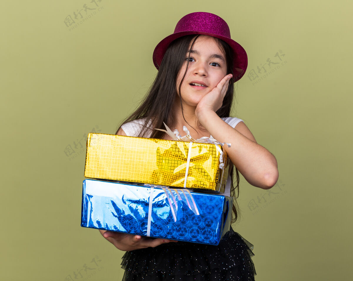 白种人惊讶的年轻白人女孩 戴着紫色派对帽 手放在脸上 拿着礼盒 隔离在橄榄绿的墙上 留着复印空间惊喜脸手