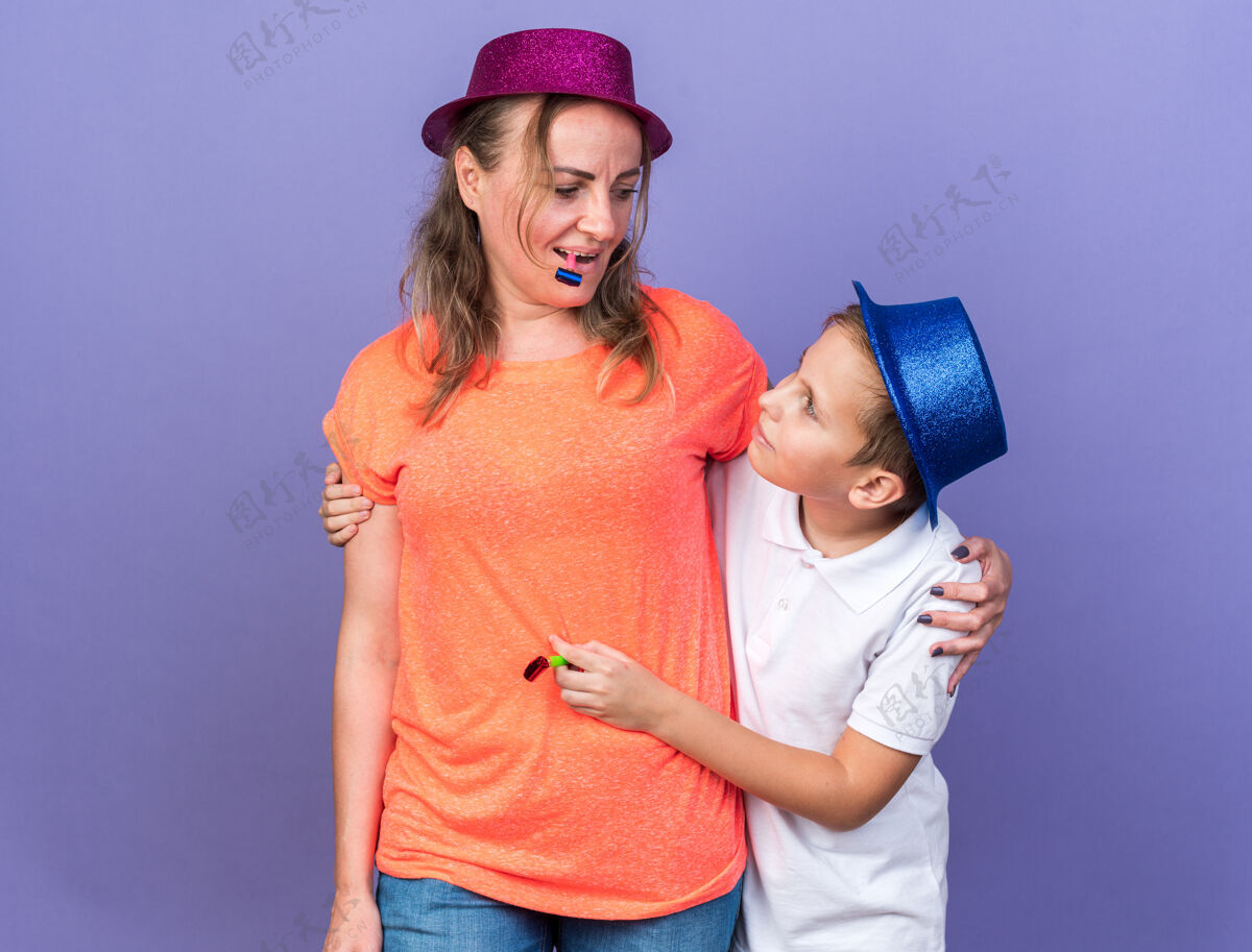 吹高兴的年轻斯拉夫男孩与蓝色党帽举行党的哨子和他的母亲站在一起 戴着紫色党的帽子和吹党的哨子孤立在紫色墙上复制空间斯拉夫人帽子年轻