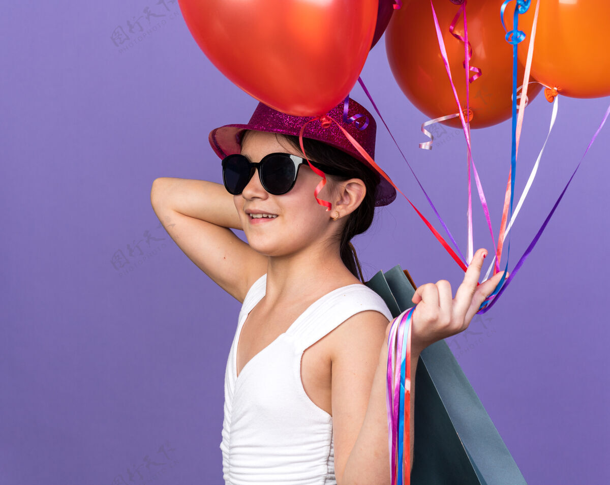 购物戴着太阳眼镜 戴着紫色派对帽 肩上扛着氦气球和购物袋 面带微笑的年轻白人女孩 隔离在紫色墙壁上 留有复印空间抱着气球白种人