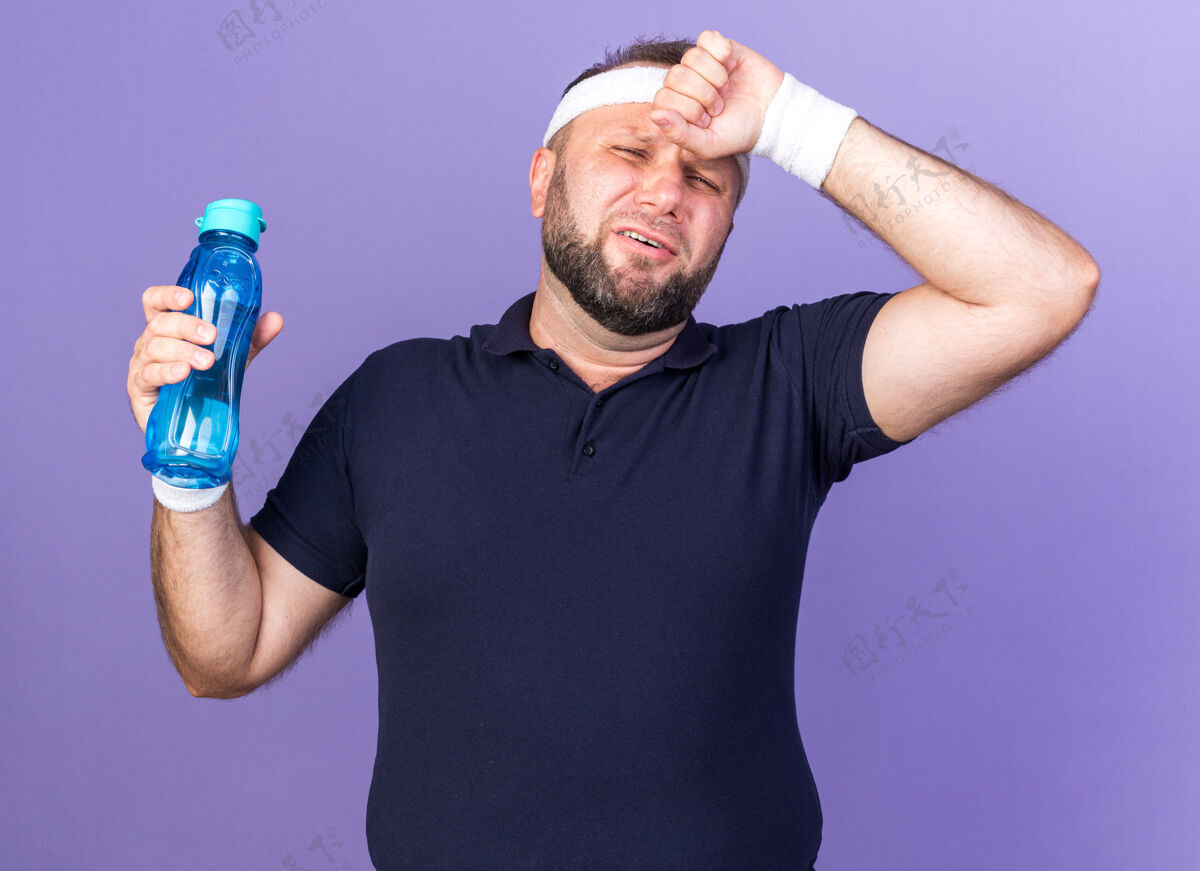 前额疼痛的成年斯拉夫运动型男子戴着头带和腕带把手放在额头上 拿着水瓶隔离在紫色的墙上 留着复制空间疼痛戴腕带