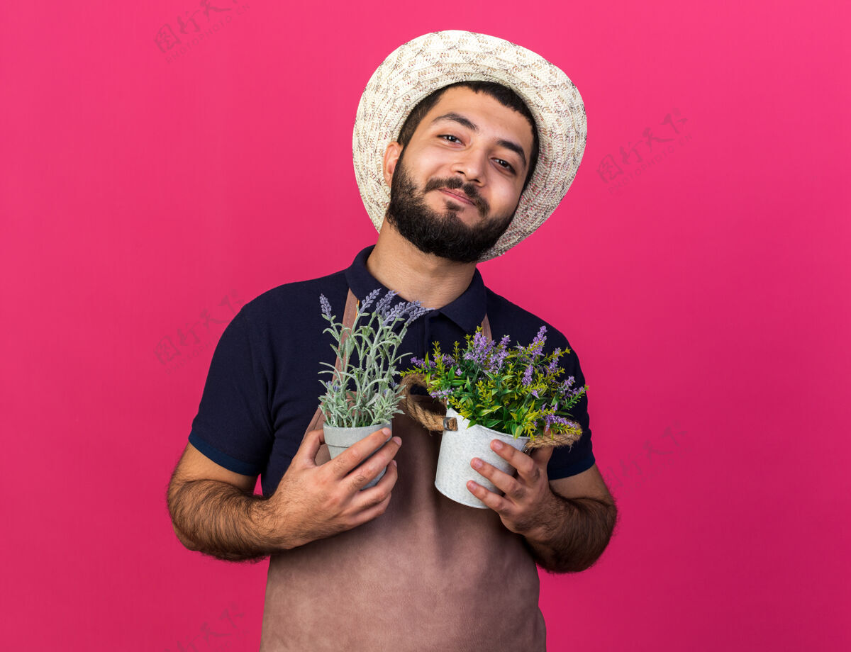请年轻的白人男园丁戴着园艺帽 手拿花盆 把花盆孤立地放在粉红色的墙上 留着复制空间年轻白种人穿