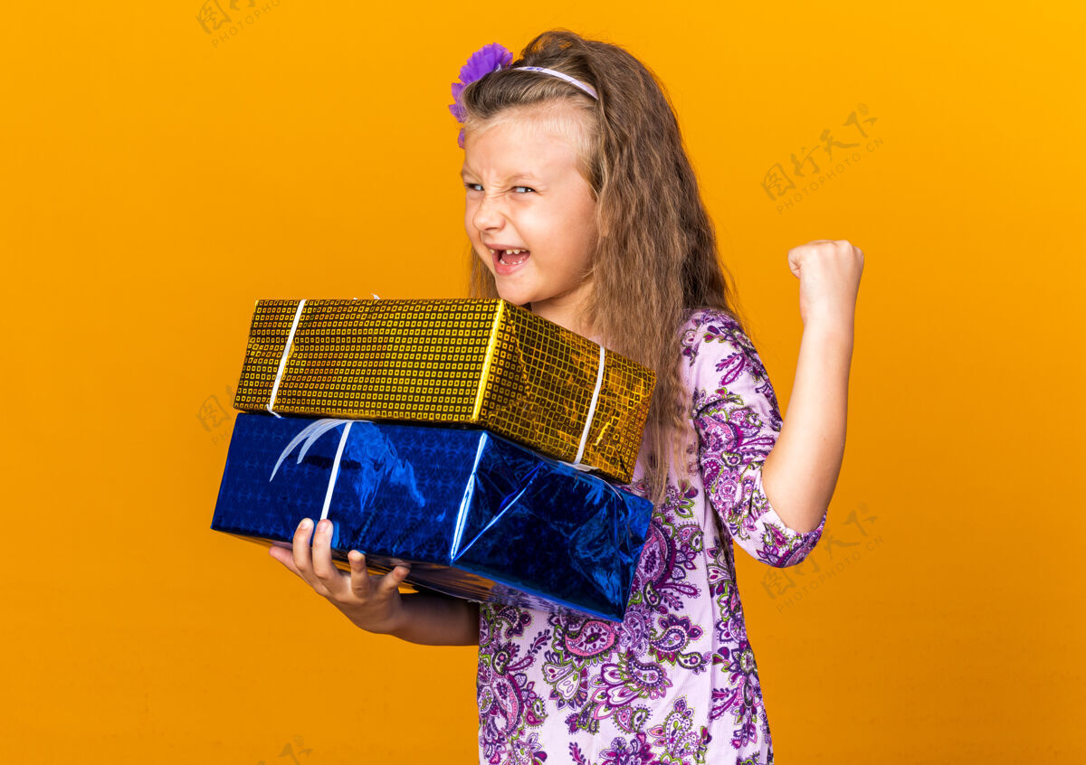 孤立快乐的小金发女孩拿着礼品盒 保持拳头在橙色的墙上与复制空间隔离抱着小拳头