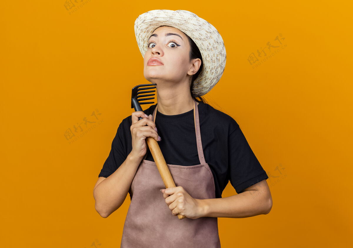 抱年轻漂亮的女园丁围着围裙 戴着帽子 拿着小耙子 站在橘色的墙上 正焦急地看着前面围裙感觉漂亮