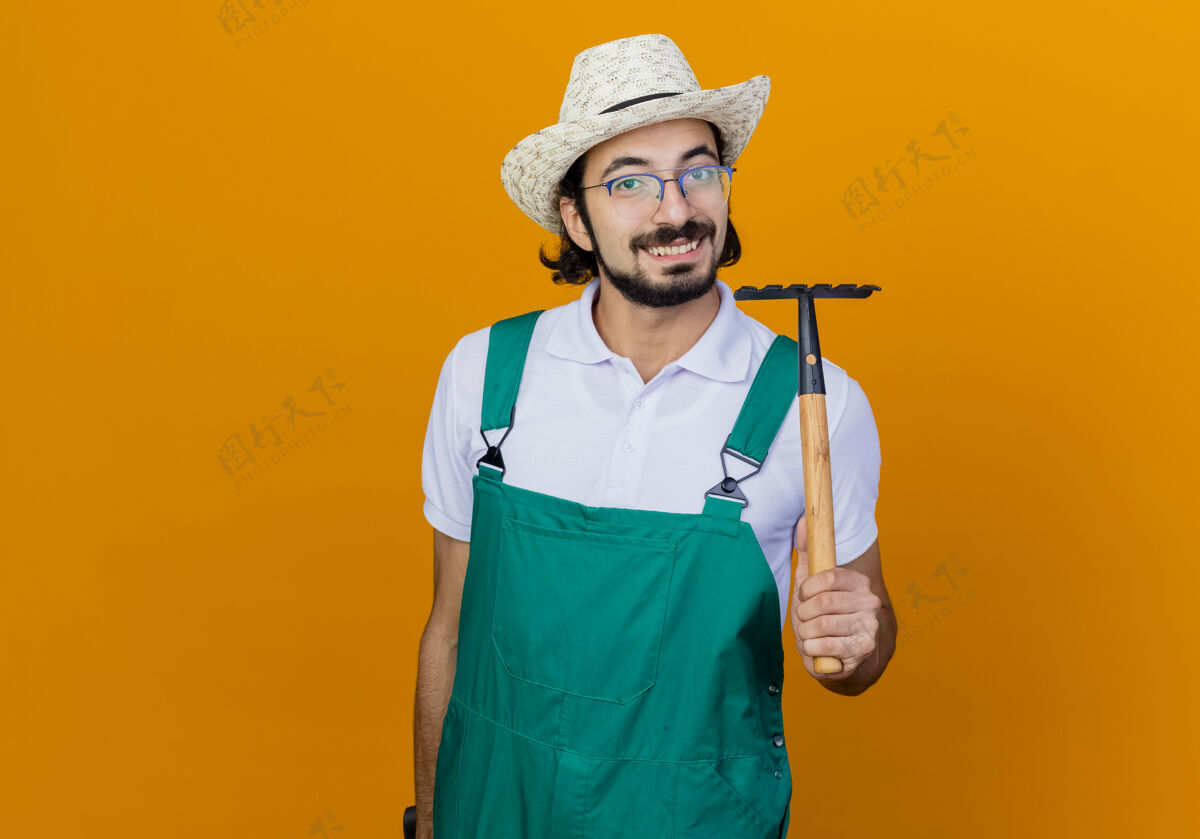 穿着年轻的留着胡须的园丁 穿着连体衣 戴着帽子 拿着小耙子 站在橙色的墙上 兴高采烈地微笑着看着前面年轻人姿势人