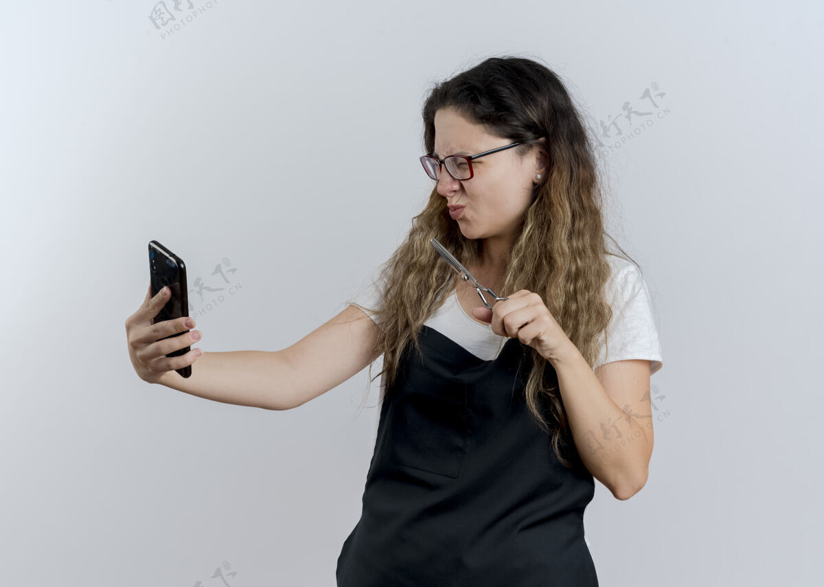 人年轻的专业美发师 围着围裙 拿着剪刀 站在白色的墙上 不高兴地看着她的智能手机屏幕专业人公民