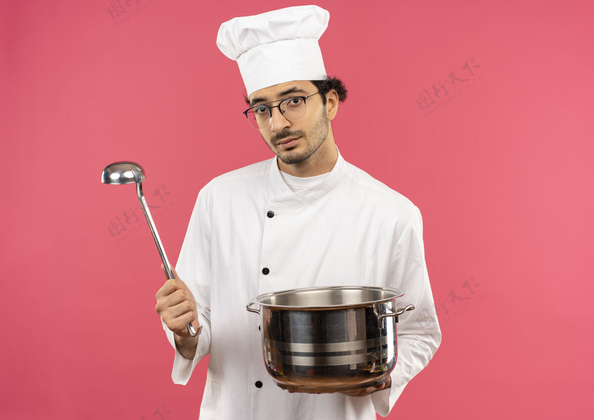 平底锅看着镜头 年轻的男厨师穿着厨师制服 戴着眼镜 手里拿着平底锅和勺子粉色年轻烹饪