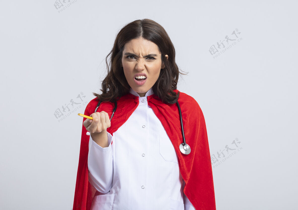 斗篷穿着医生制服 戴着红色斗篷和听诊器的笨拙女超人拿着铅笔 看着隔离在白墙上的前面姿势脸女孩