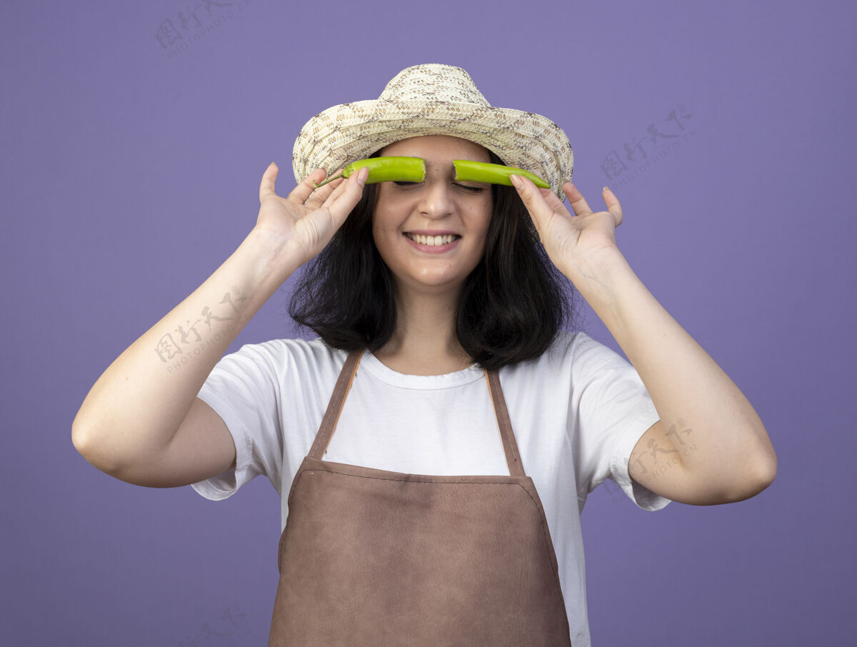 眼睛年轻的黑发女园丁穿着制服 戴着园艺帽 眼睛上涂着半个辣椒 隔离在紫色的墙上一半穿市民