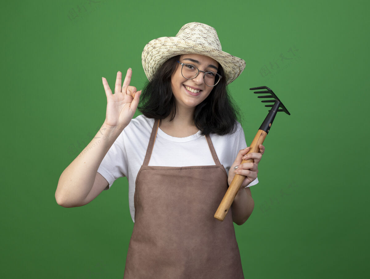 光学微笑着的年轻黑发女园丁戴着眼镜和制服 戴着园艺帽 打着ok手势 拿着绿色墙上的耙子眼镜手拿着