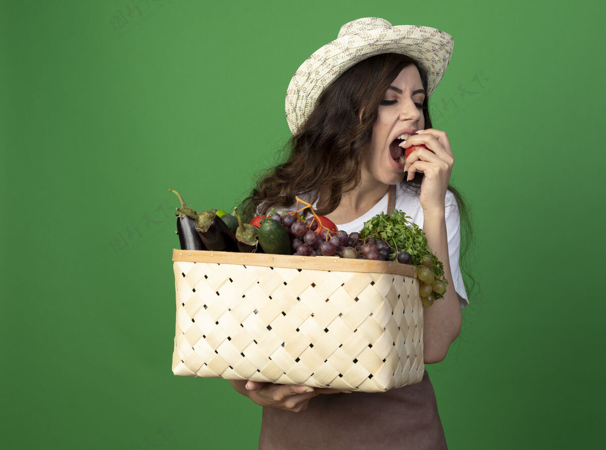 人饥饿的年轻女园丁穿着制服 戴着园艺帽 拿着菜篮子 假装咬隔离在绿墙上的西红柿人西红柿园艺