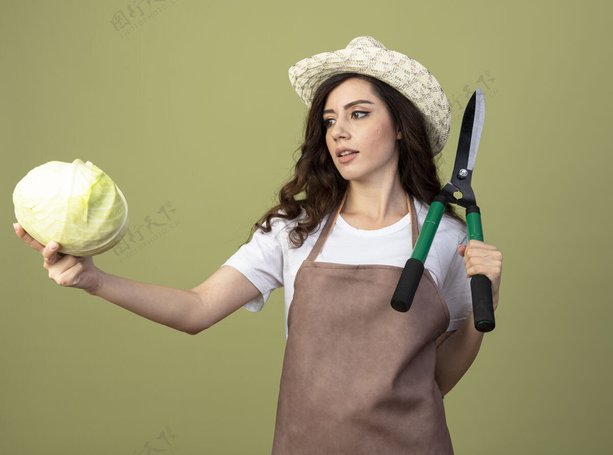 表情自信的年轻女园丁穿着制服 戴着园艺帽 拿着园艺剪 看着橄榄绿墙上孤立的卷心菜帽子自信橄榄