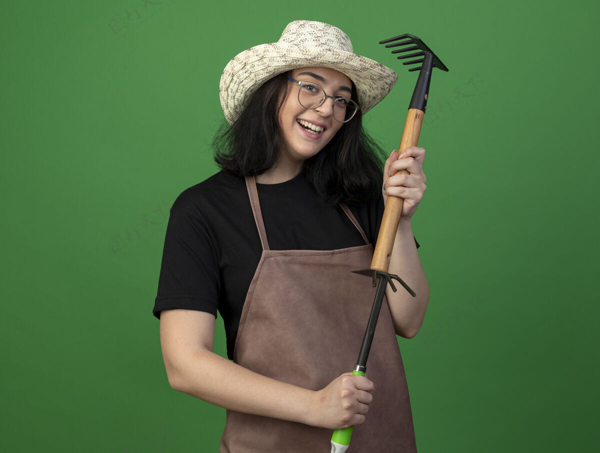 姿势微笑着的年轻黑发女园丁戴着眼镜和制服戴着园艺帽拿着耙子锄头耙子隔离在绿色的墙上表情市民耙子