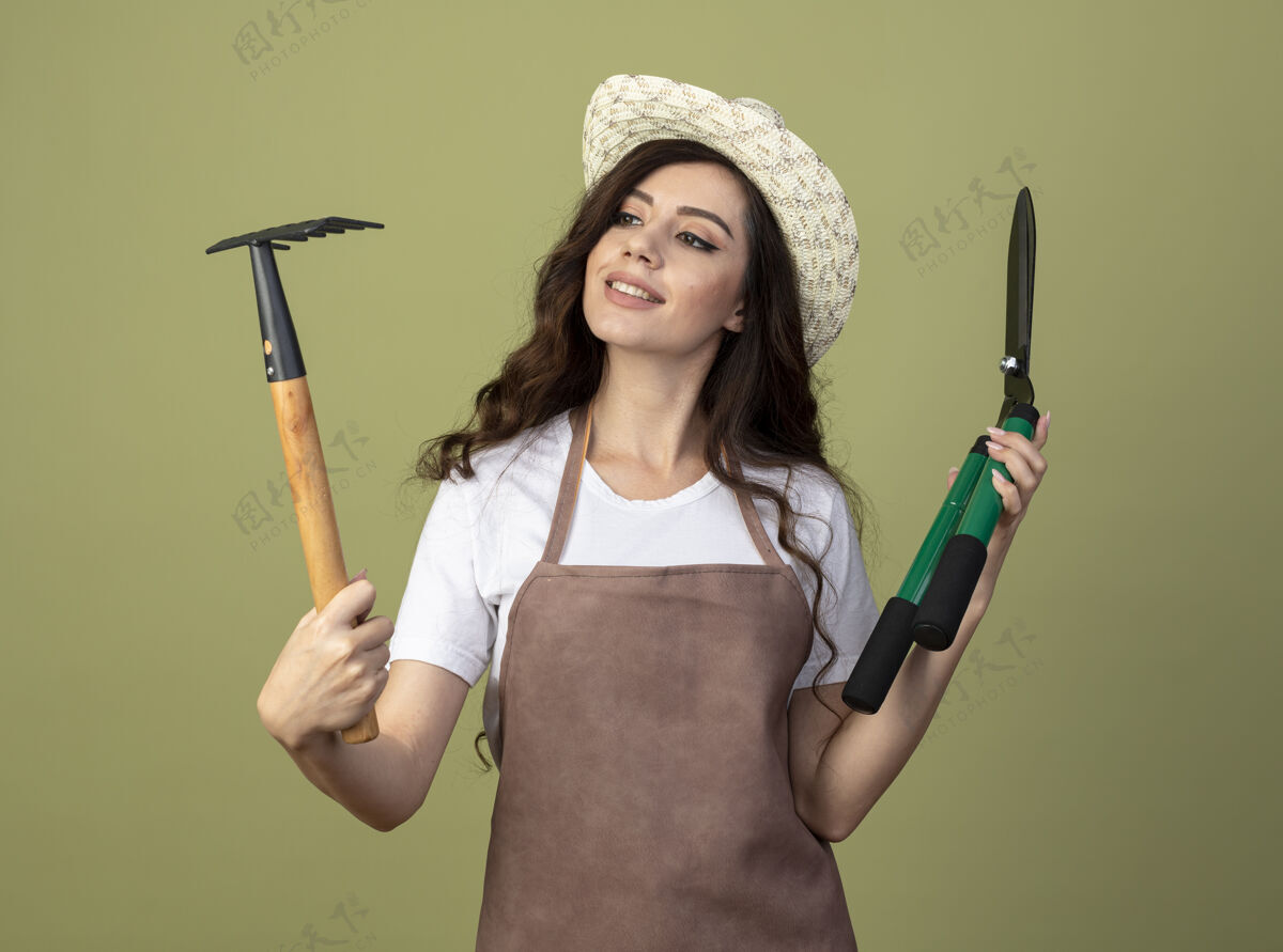 年轻人身着制服 头戴园艺帽 面带微笑的年轻女园丁手持剪刀 看着橄榄绿墙上孤立的耙子表情持有制服