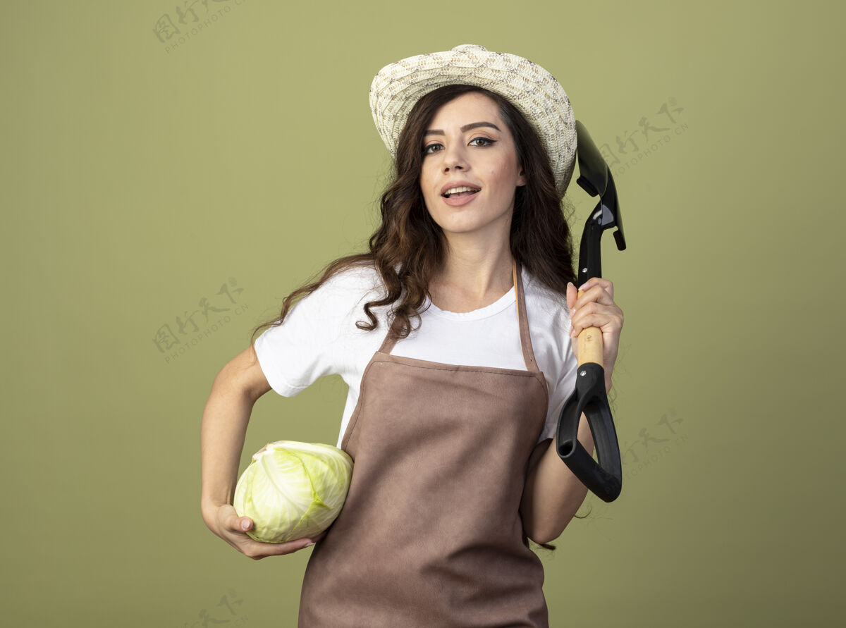 橄榄自信的年轻女园丁穿着制服 戴着园艺帽 把卷心菜和铲子孤立地放在橄榄绿的墙上举行人自信