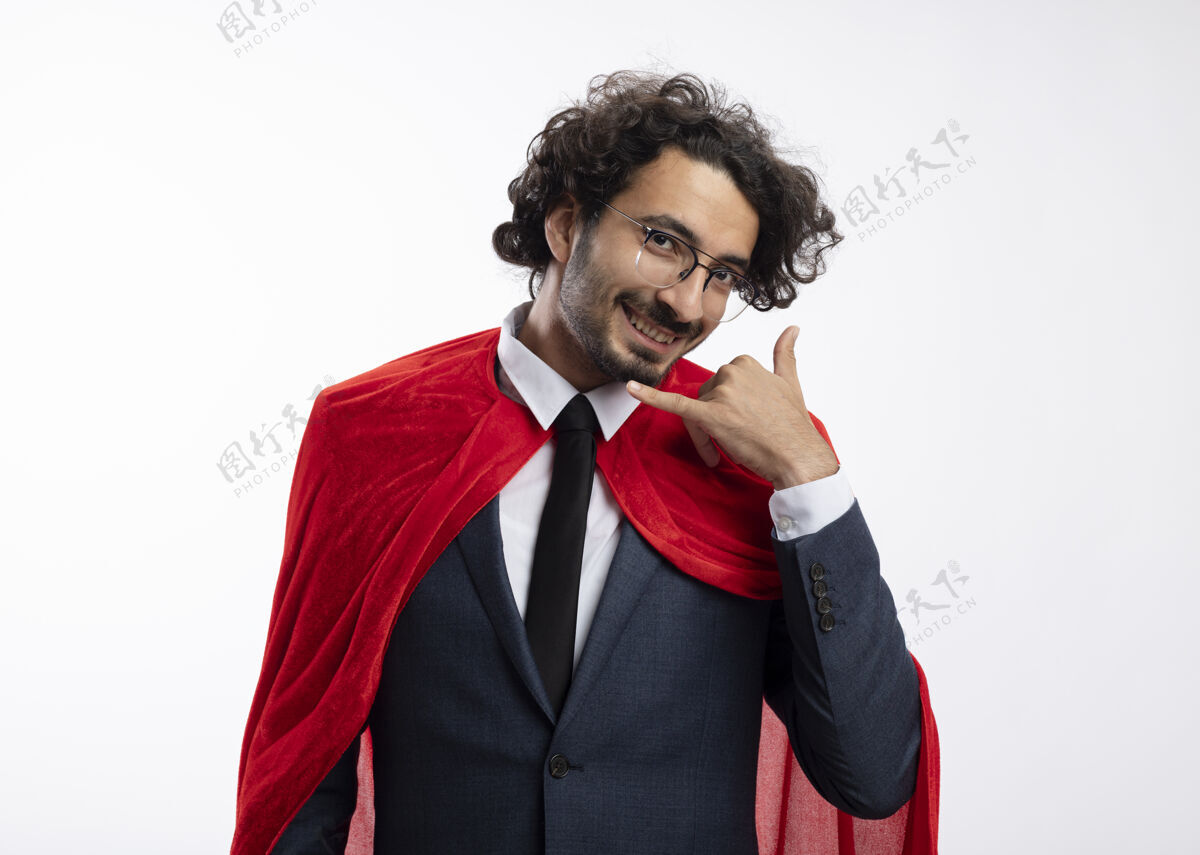 人微笑着的年轻超级英雄 戴着眼镜 穿着西装 披着红色斗篷 在白色的墙上打着“叫我”的手势套装表情人