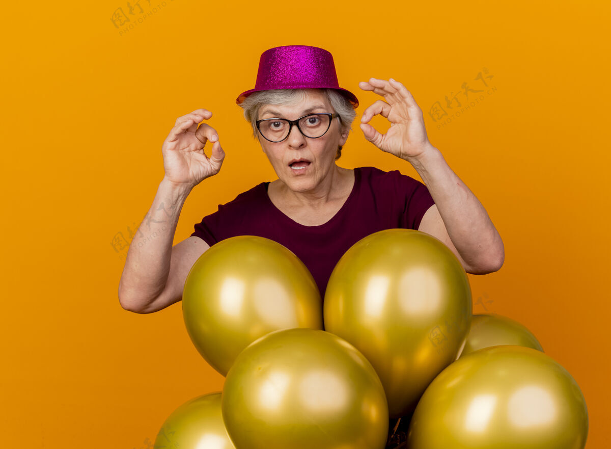 女人一位戴着光学眼镜 戴着派对帽 手持氦气球的老妇人站在那里 两手孤立在橙色的墙上 做着“okhand”的手势手眼镜穿