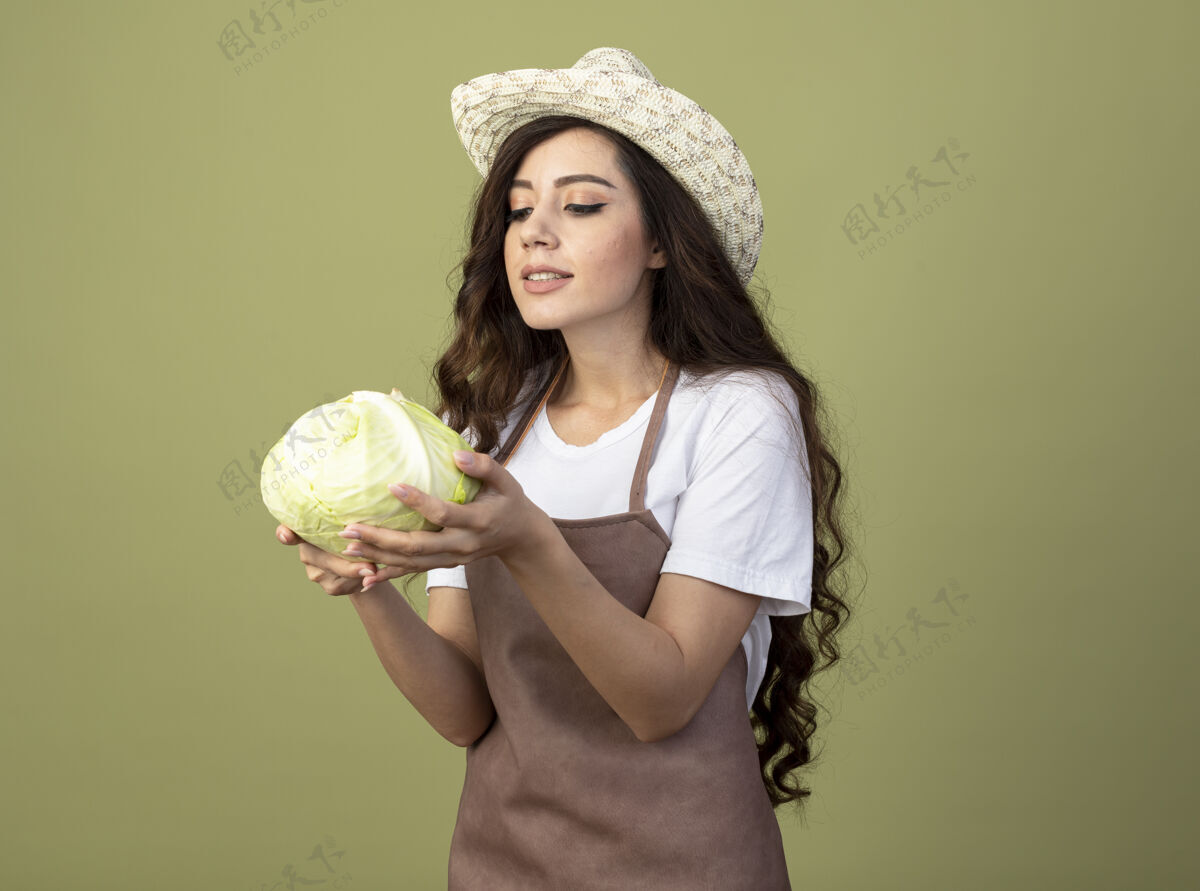 制服自信的年轻女园丁穿着制服 戴着园艺帽 站在橄榄绿的墙上看着孤立的卷心菜女人年轻姿势