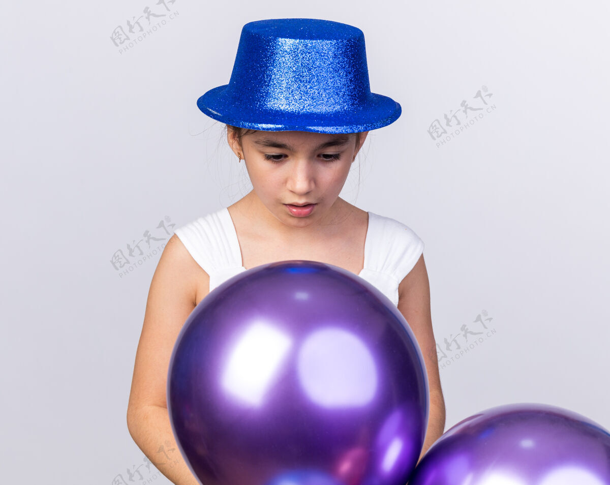年轻兴奋的年轻白种人女孩戴着蓝色的派对帽 看着隔离在白色墙壁上的氦气球 还有复制空间兴奋氦女孩