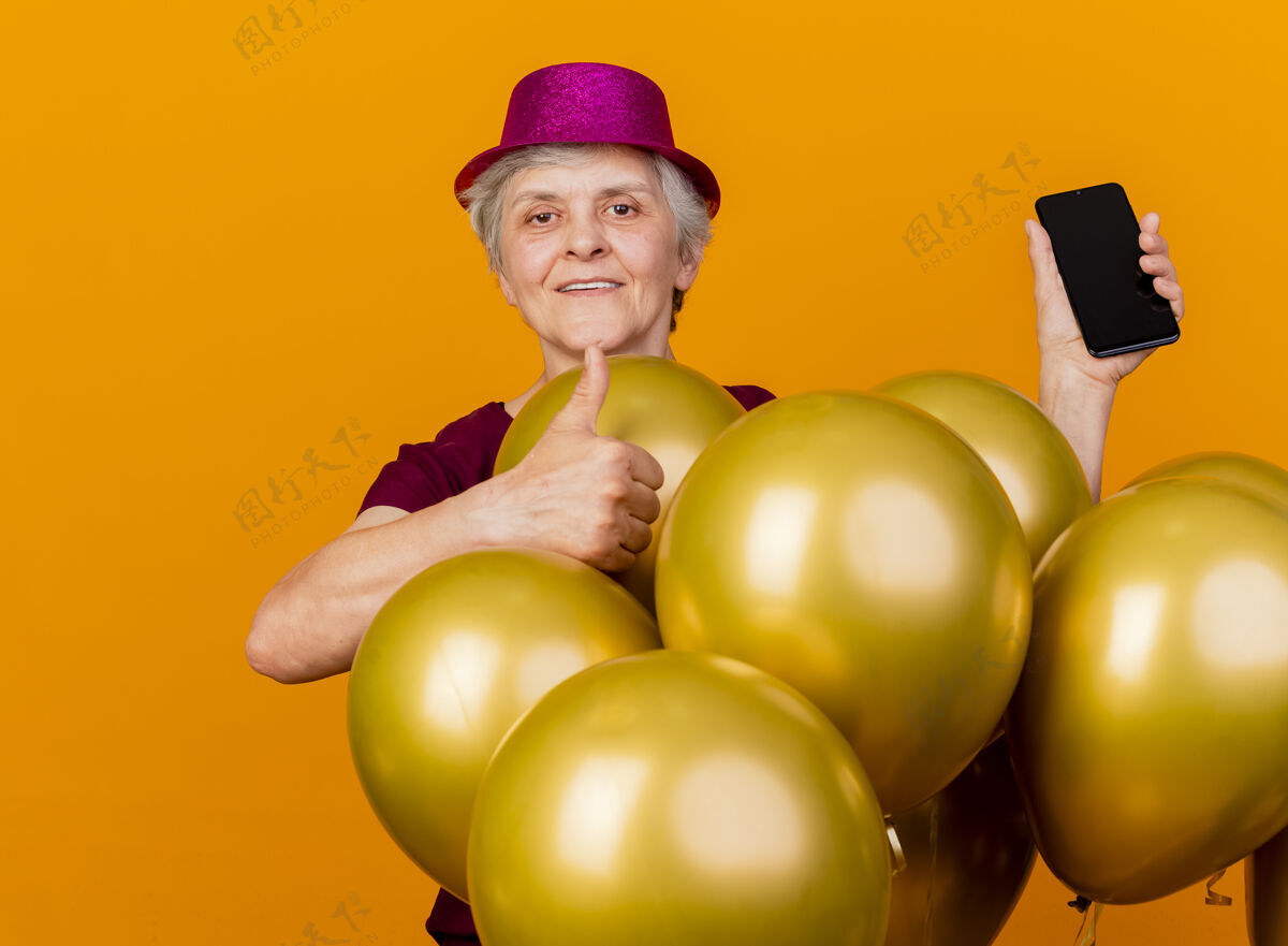 氦戴着派对帽的老太太高兴地竖起大拇指 站在橙色的墙上孤立地拿着电话站市民人