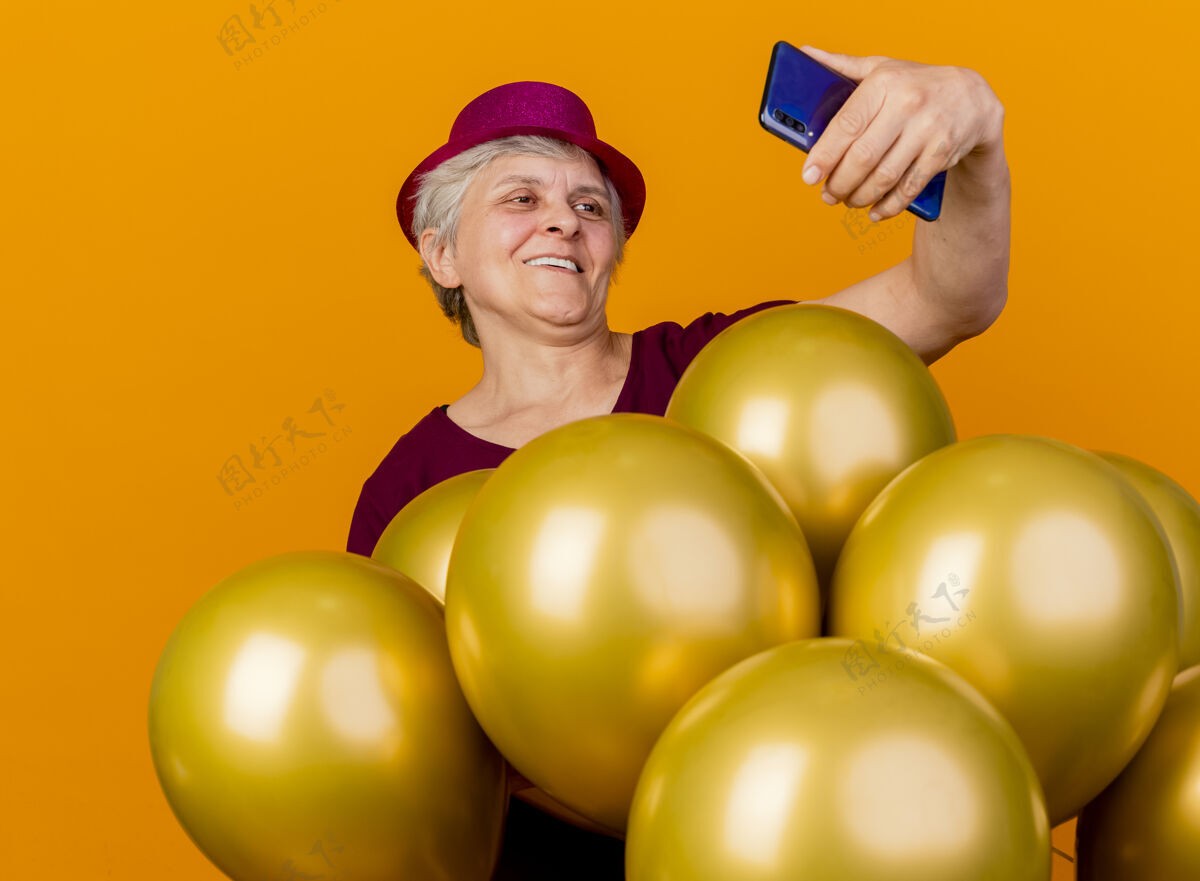帽子微笑着戴着派对帽的老妇人站在氦气球旁 看着隔离在橙色墙上的手机市民姿势站