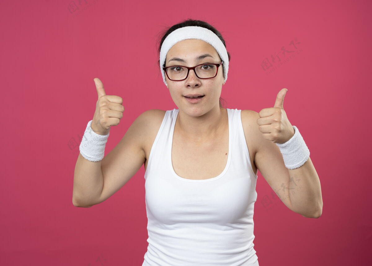 年轻人这位戴着眼镜 戴着头带和腕带的年轻运动女性给人留下了深刻印象 她竖起大拇指 两只手分别放在粉红色的墙上拇指头带戴