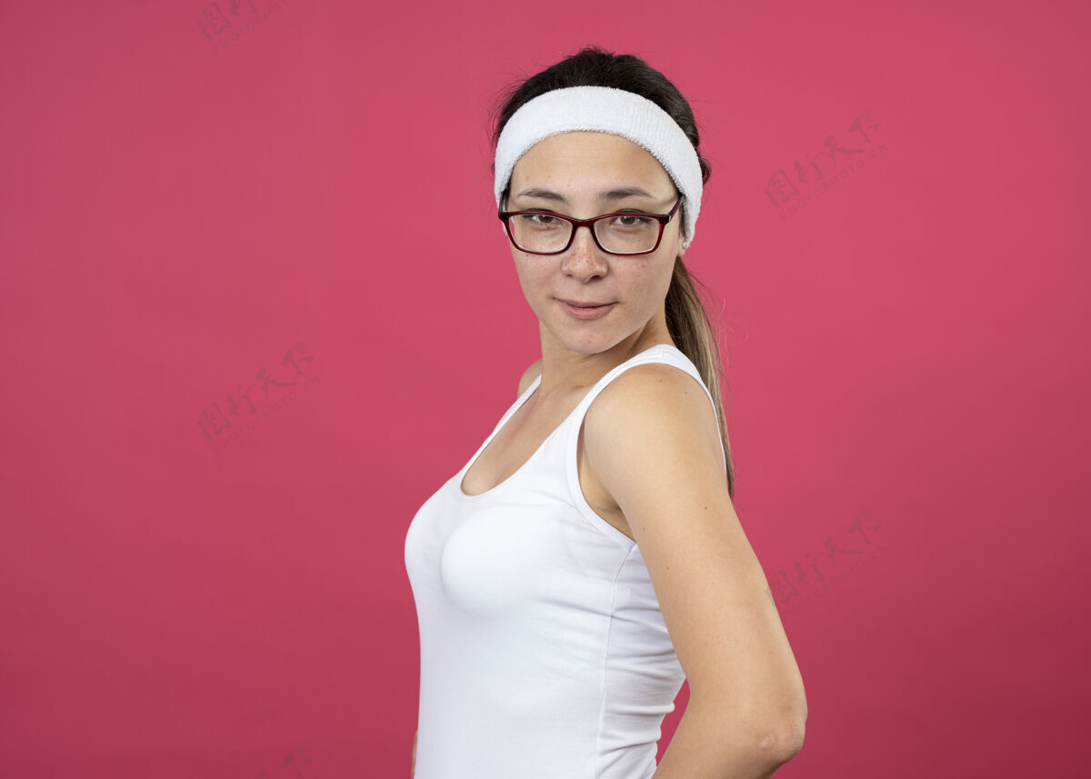 女人戴着眼镜 戴着头带和腕带的快乐的年轻运动女性站在粉红色的墙上 与外界隔绝腕带女孩年轻