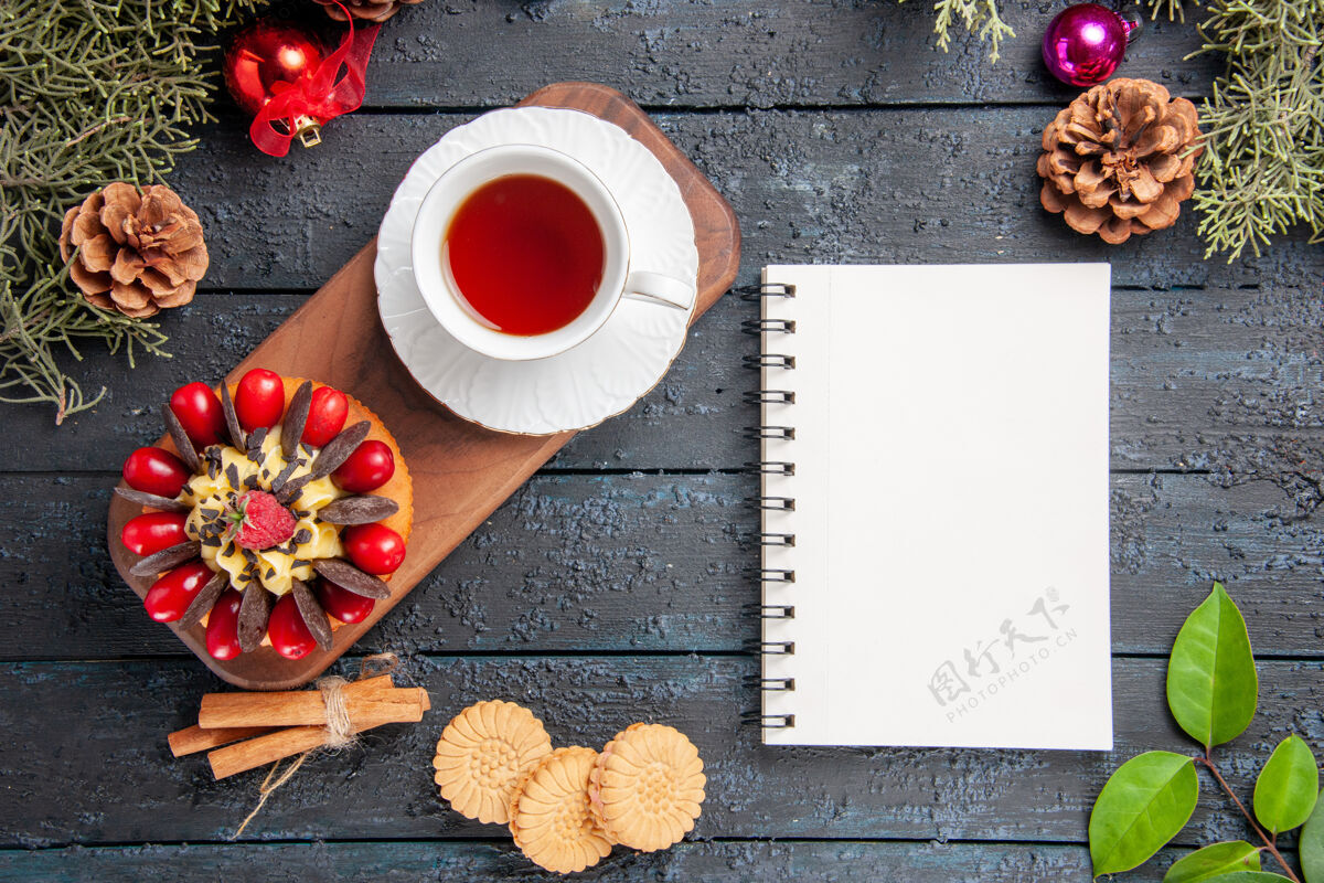 松果顶视图一杯茶和浆果蛋糕放在木制盘子上松果圣诞玩具叶子和一个笔记本放在深色的木制桌子上木头深色的茶