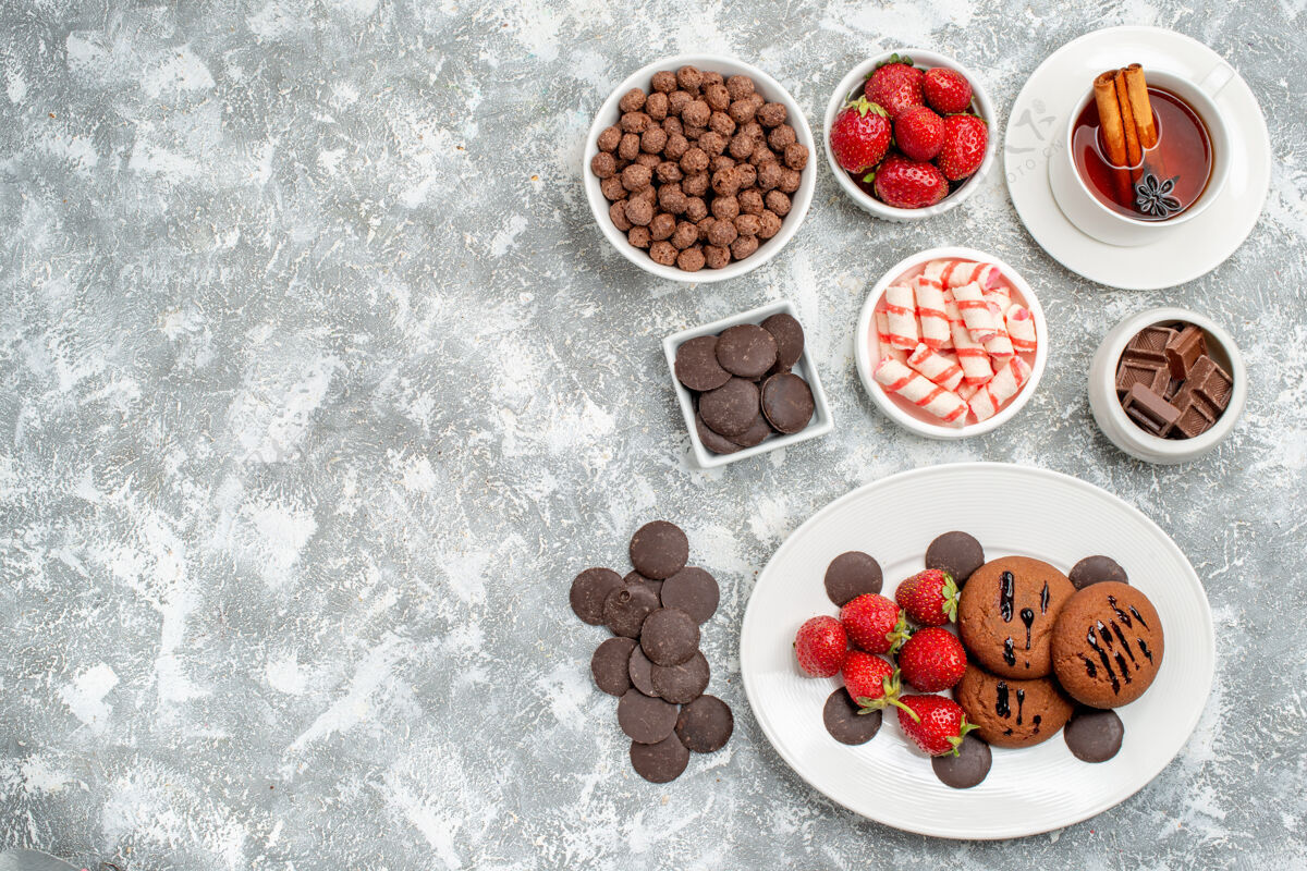杯子顶视图饼干草莓和圆形巧克力放在椭圆形盘子碗上 糖果草莓和巧克力麦片和一杯茶放在灰白色桌子的右侧 有自由空间谷类食品盘子草莓