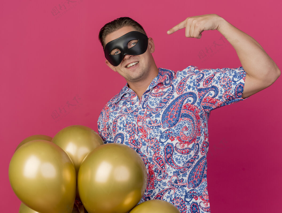 粉色面带微笑的年轻人戴着化装眼罩拿着气球指着自己孤立的粉红色他自己化装舞会气球
