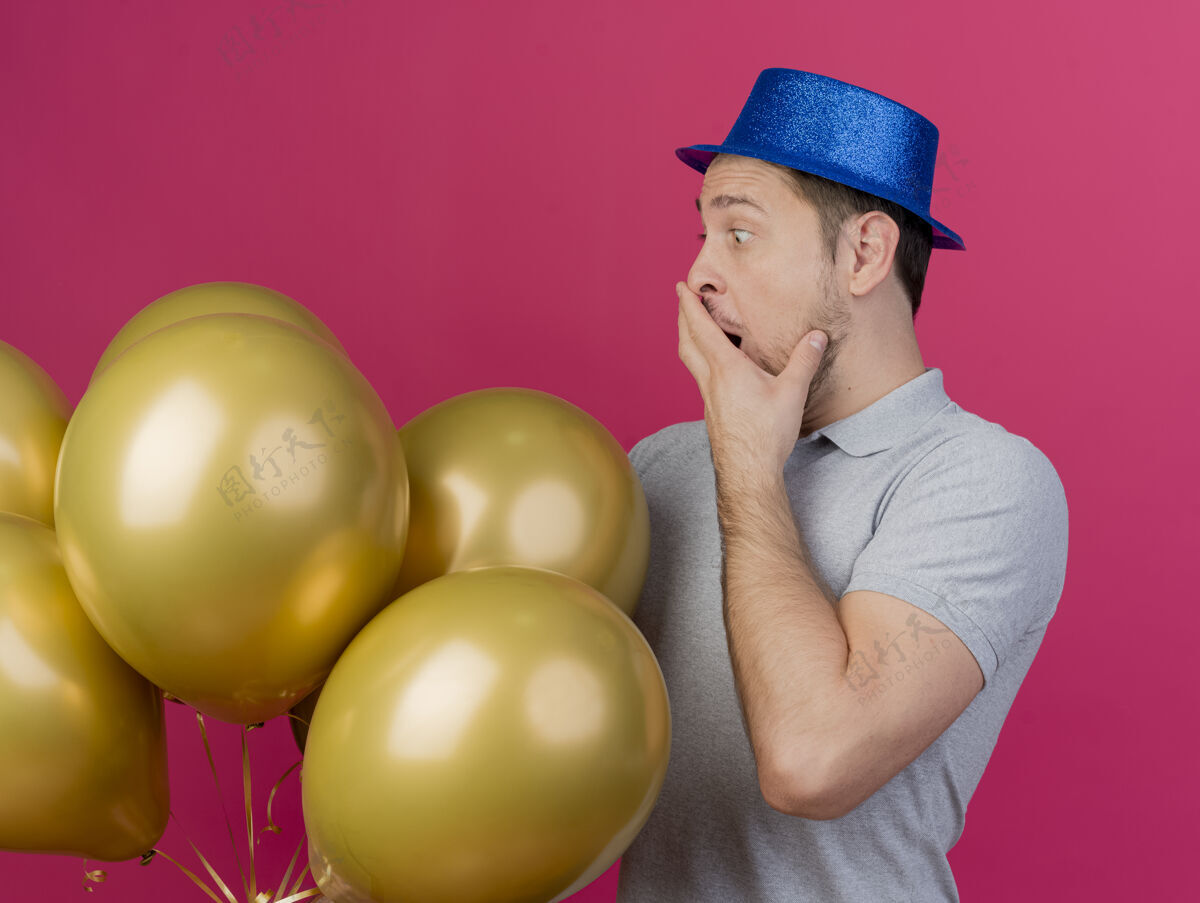 派对惊讶的年轻人戴着蓝色的派对帽 拿着气球 看着满嘴都是粉红色孤立的手蓝色粉色帽子
