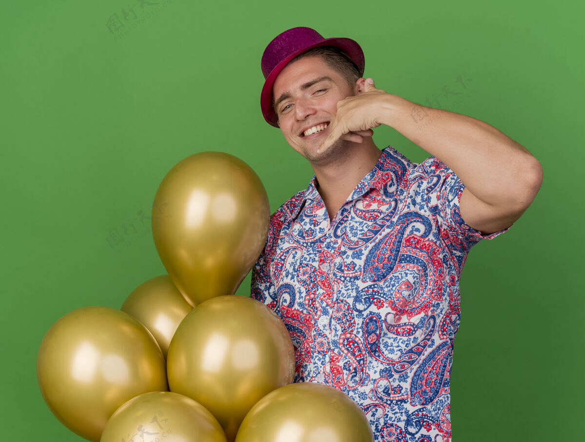帽子微笑着的年轻人戴着粉红色的帽子 拿着气球 在绿色的草坪上展示着打电话的手势绿色穿举行