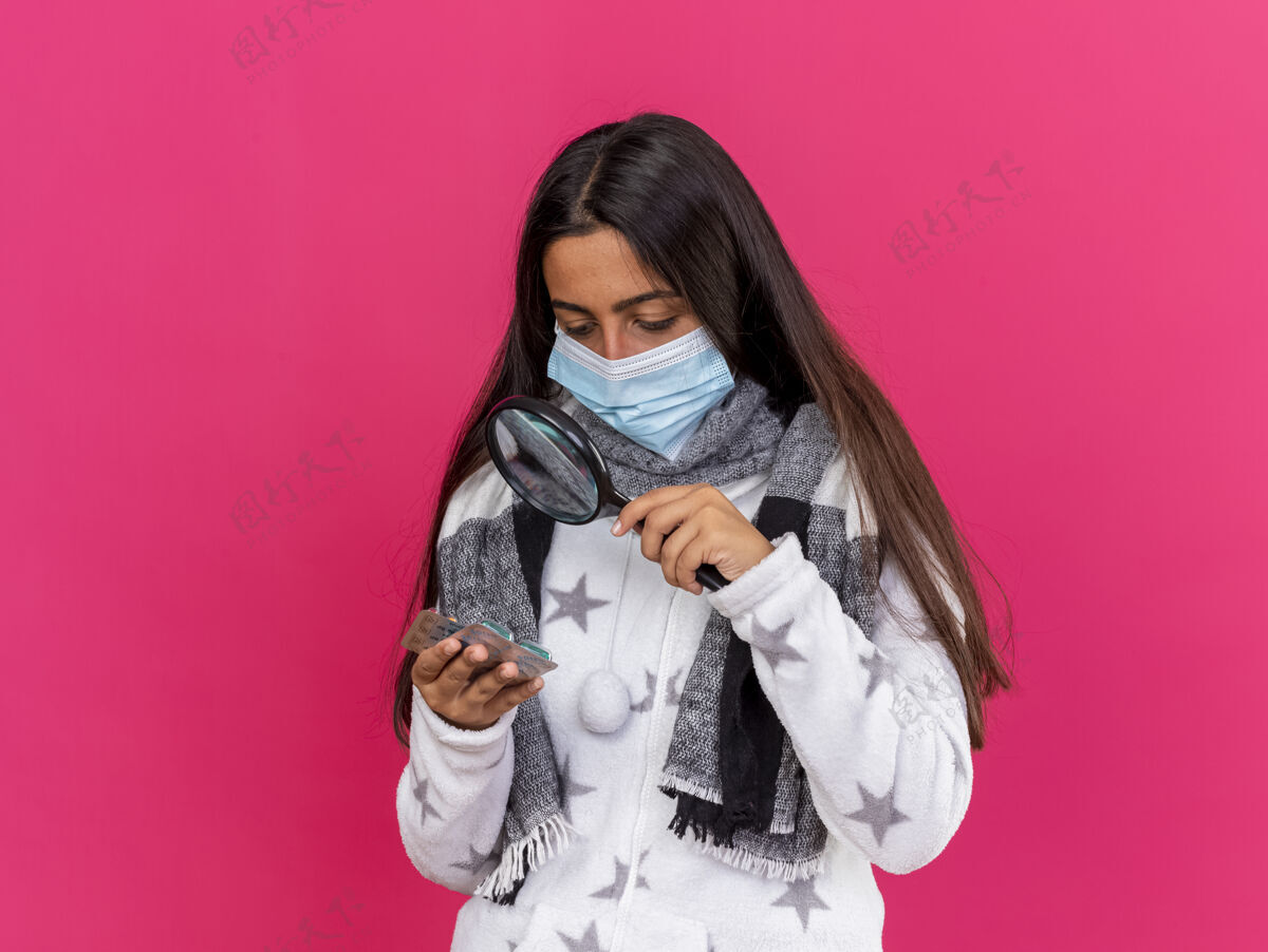 面具年轻的生病的女孩戴着医用面罩 戴着围巾 拿着放大镜看着粉红色的药片穿年轻持有