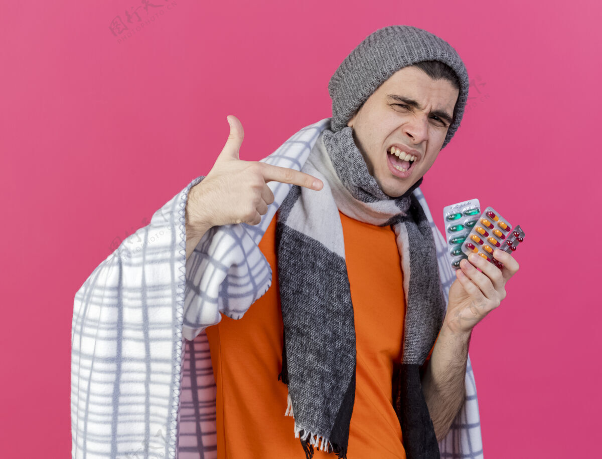 穿一个戴着冬天帽子 围巾裹着格子布的快乐的生病的年轻人 指着粉红色的药片男人喜悦年轻