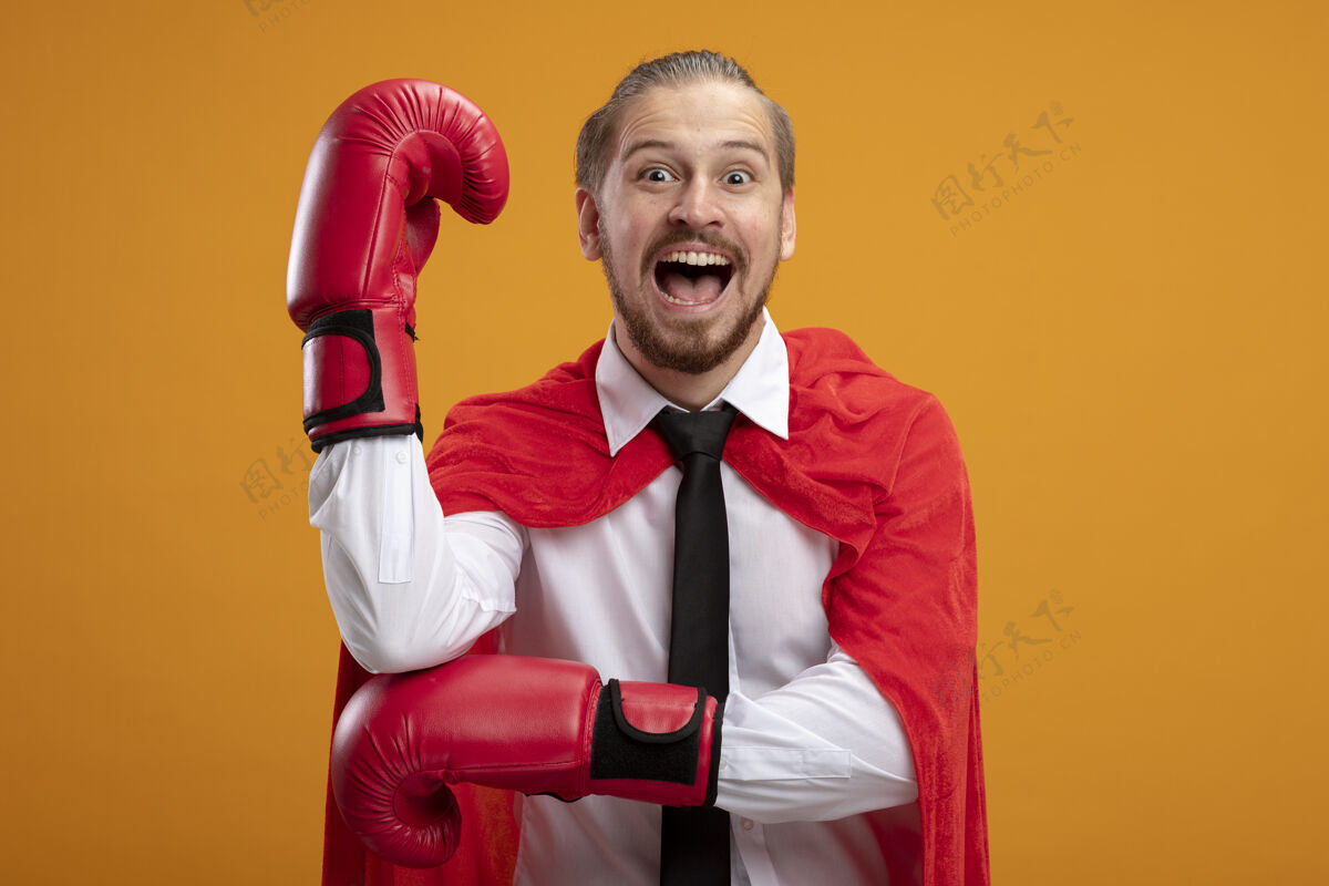 戴上快乐的年轻超级英雄戴着领带和拳击手套举起拳头孤立在橙色拳击拳头橙色
