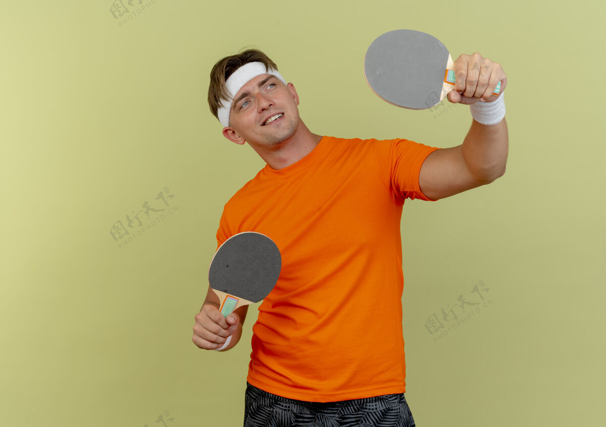 乒乓球面带微笑的年轻英俊的运动型男子戴着头带和腕带举着乒乓球拍 看着橄榄绿墙上孤立的一面腕带人绿色