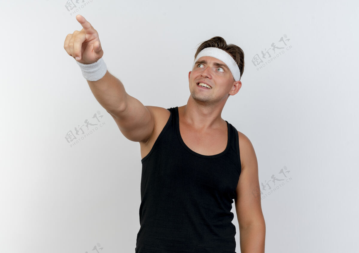 腕带面带微笑的年轻英俊的运动型男子戴着头带和腕带 抬头指着隔离在白墙上的一侧指点姿势头带