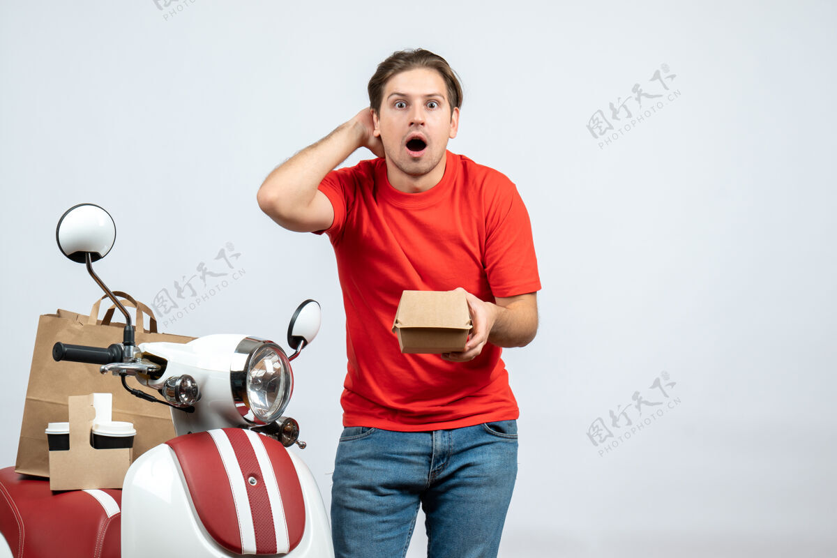 抱身穿红色制服的送货员站在滑板车旁 手里拿着白色背景的小盒子 吓了一跳男盒子近