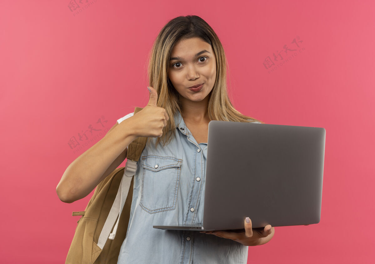 女孩高兴的年轻漂亮的女学生穿着背包拿着笔记本电脑 并显示拇指孤立在粉红色的墙上粉色表情穿着