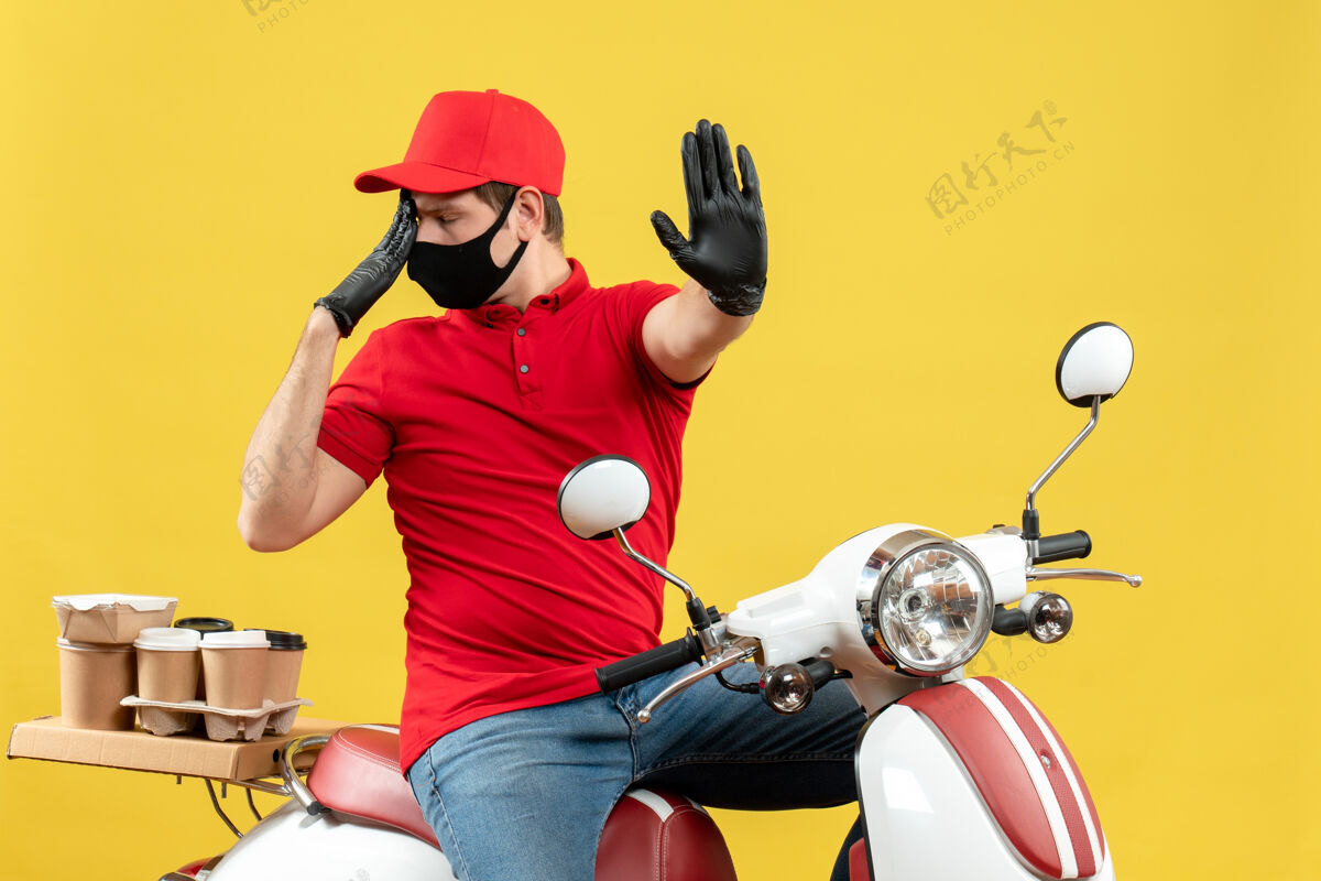 球前视图情绪化的年轻人穿着红色上衣 戴着帽子手套 戴着医用面罩 坐在黄色背景的踏板车上传递命令男子命令车辆