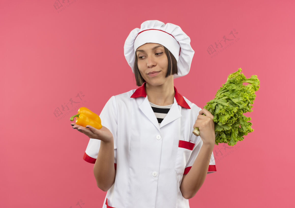 粉红身着厨师制服的年轻女厨师手拿胡椒和莴苣 看着粉红色墙上孤立的胡椒人抱人