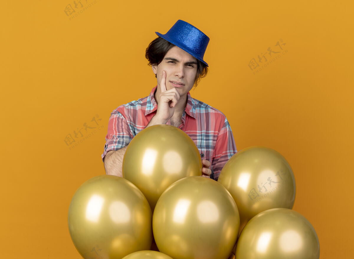 人戴着蓝色派对帽的迷茫帅哥把手放在下巴上 站在橙色墙上孤立着氦气球穿困惑公民
