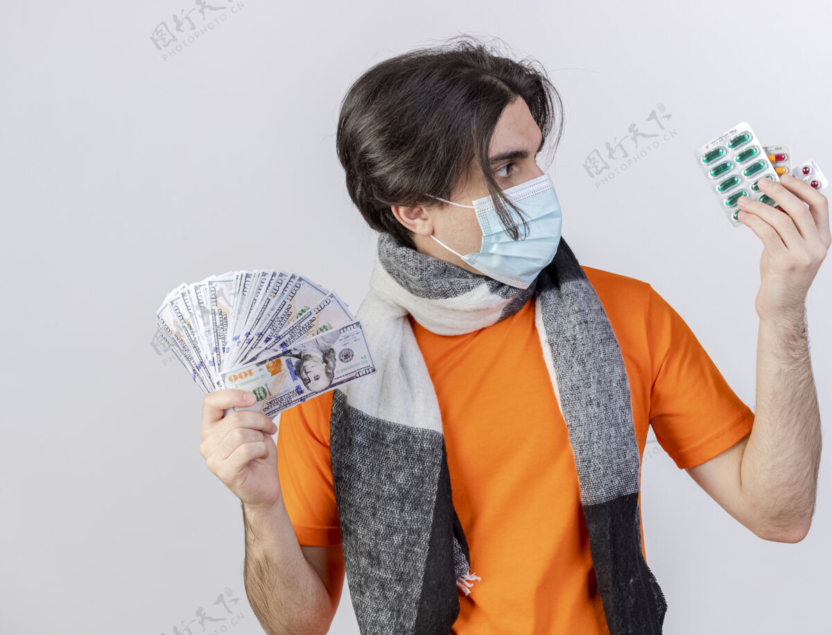 男人年轻的病夫戴着围巾和医用口罩 手里拿着现金 看着药片上的白色隔离物医疗疾病持有