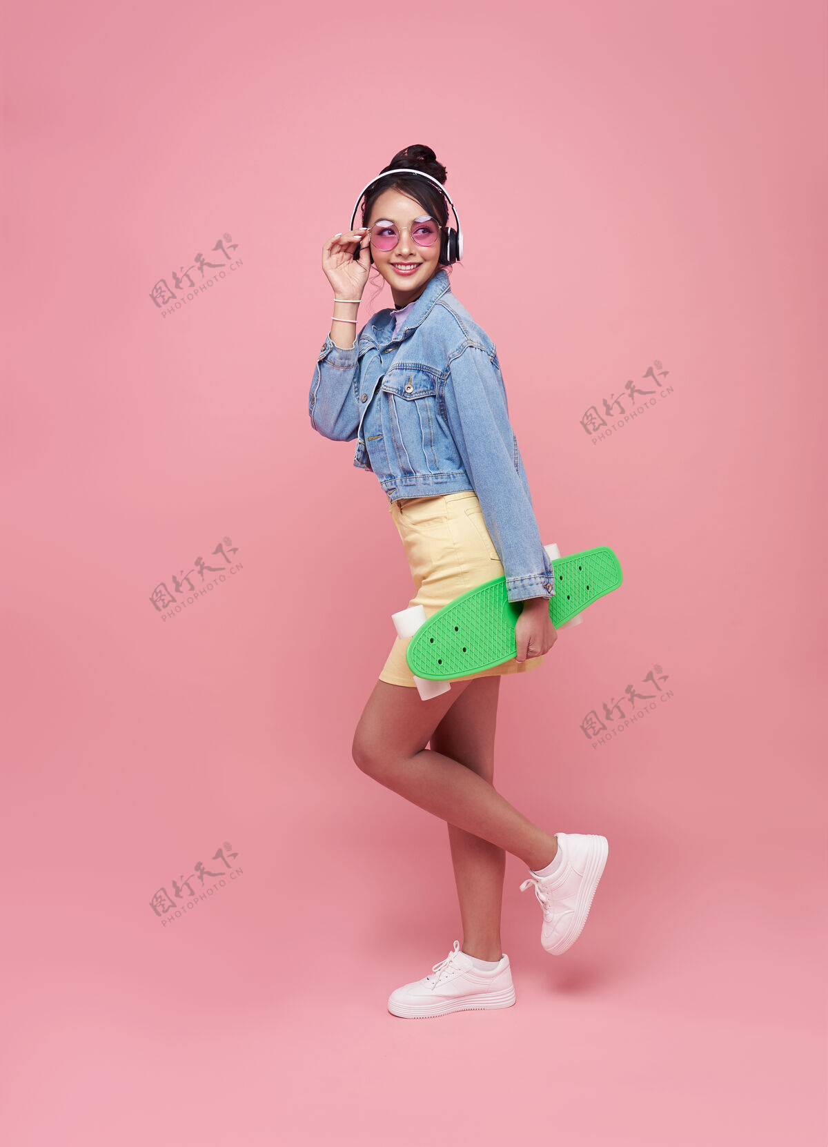 乐趣年轻的亚洲少女手持滑板 戴着无线耳机 在粉红色的墙上听音乐微笑情感快乐
